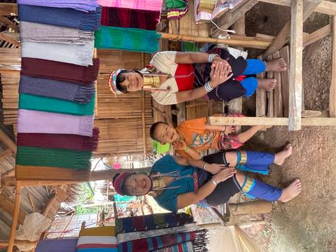       Three people in traditional attire sitting on a wooden bench under colorful hanging textiles.
  