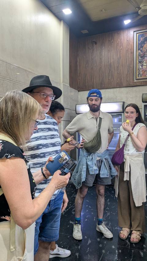       Group of tourists indoors, some holding phones, one eating a snack.
  
