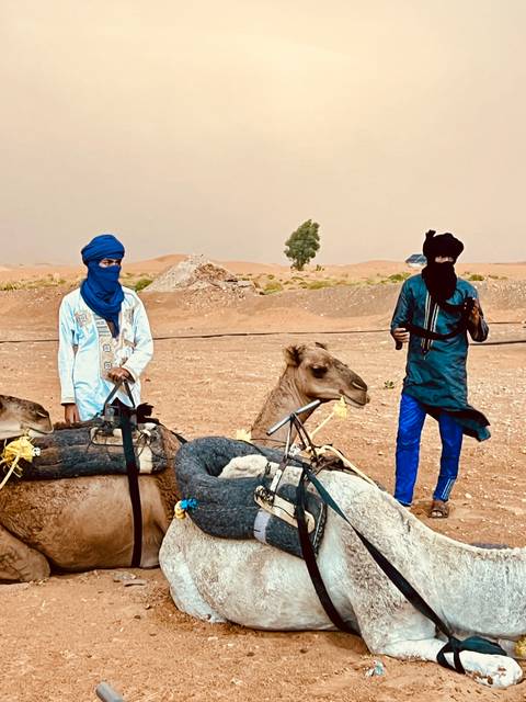 Two people in traditional attire with camels in a desert landscape.