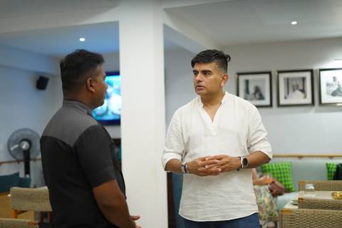       Two men engaged in a conversation inside a room with framed pictures.
  