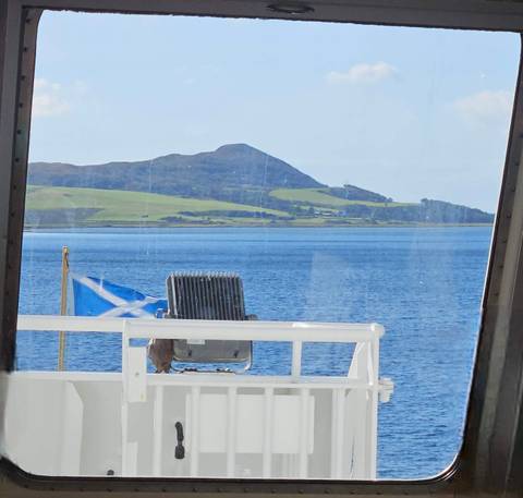       View from a boat with a Scottish flag towards a hilly landscape.
  