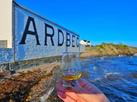       Glass of whisky held up in front of the Ardbeg sign by the water.
  