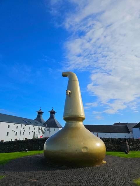       Large sculpture of a distillery column against a blue sky with roofs visible.
  