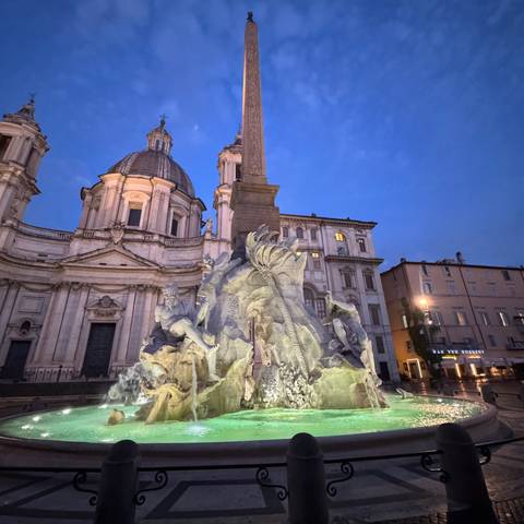       A magnificent fountain with statues and an obelisk in a lit-up square during early evening.
  