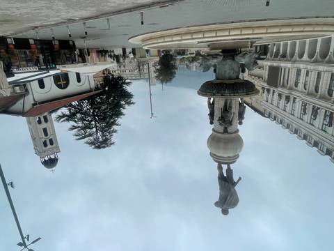       Statue and fountain in a city square with historic buildings, overcast sky.
  