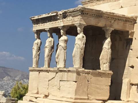       The Caryatids of the Erechtheion on the Acropolis, with city and mountain in the background.
  