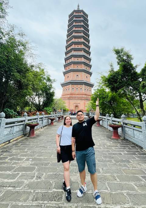       Couple standing in front of a traditional red tower surrounded by greenery.
  