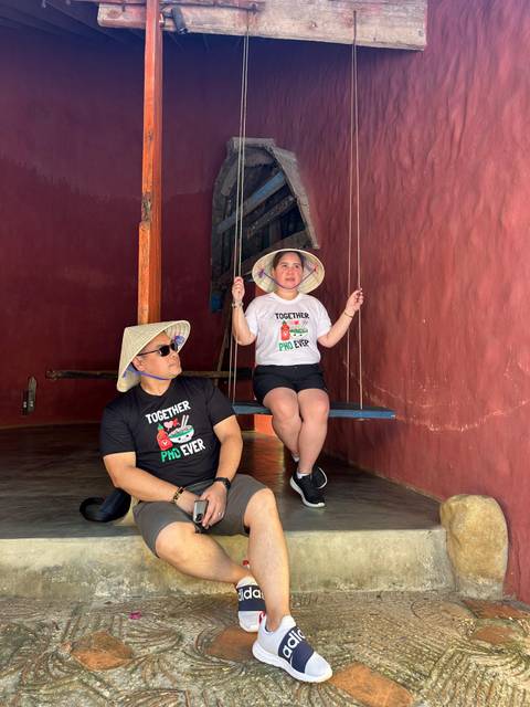       Two people on a swing inside a red-walled room wearing Vietnamese hats.
  