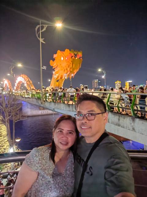       Couple taking a selfie on a bridge at night with crowd and city lights.
  