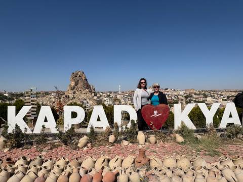       Two women posing in front of a Cappadocia sign with rock formations in the background.
  