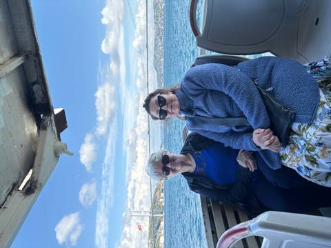       Two people sitting on a boat with water and a bridge in the background, under a partly cloudy sky.
  