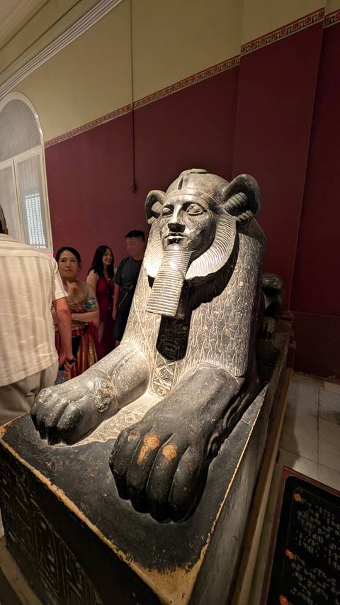       Ancient Egyptian statue with people taking photos around it in a museum setting.
  
