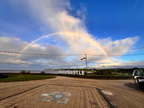       A rainbow over a scenic area with a 'Ballycastle' sign and hills in the background.
  