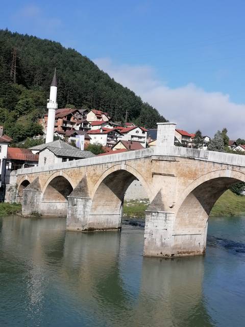       Historic stone bridge over a river with village in the background.
  