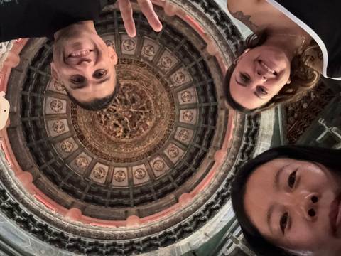       Three people posing under a dome with intricate ceiling.
  