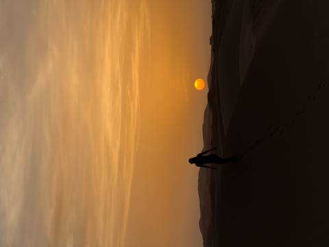       Silhouette of a person walking on sand dunes at sunset.
  