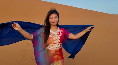 A woman holding a blue scarf with dunes in the background.