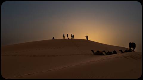 Silhouettes of people and camels on a sand dune at sunset.