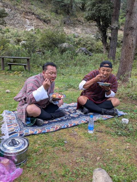       Two men sitting on a rug eating traditional food outdoors.
  