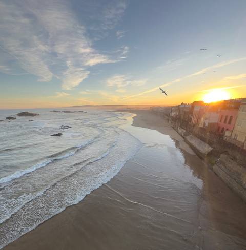       Sunset over a coastal beach with waves and cityscape.
  