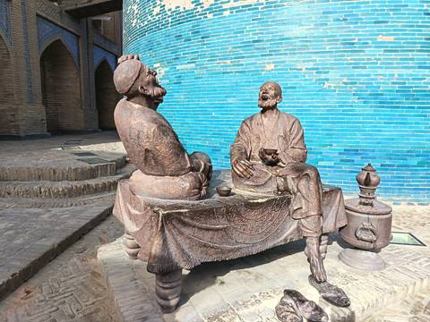 Bronze statue of two men sitting at a table by a blue wall.
