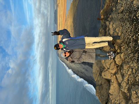 A couple posing on a cliff with a view of a black sand beach and ocean.