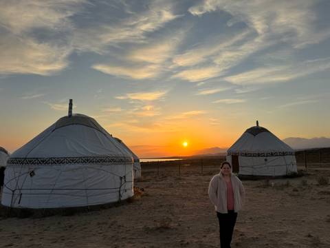       A person smiling in front of a row of yurts at sunset.
  