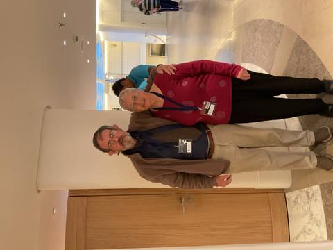 Two people posing in an interior setting with other people in the background.