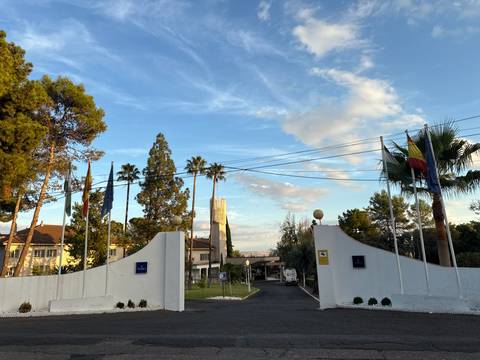 Entrance gate to a large property with flagpoles and palm trees.