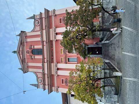       Pink facade of a church with trees and people around.
  