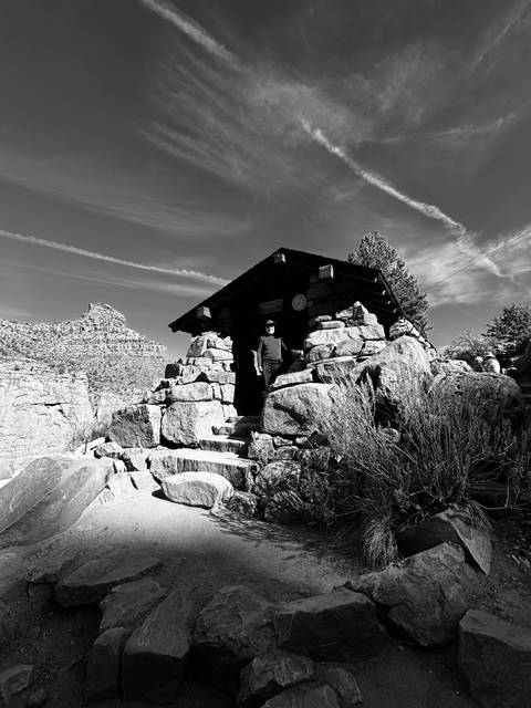       Black and white photo of a person standing at a small stone building on a mountain trail.
  