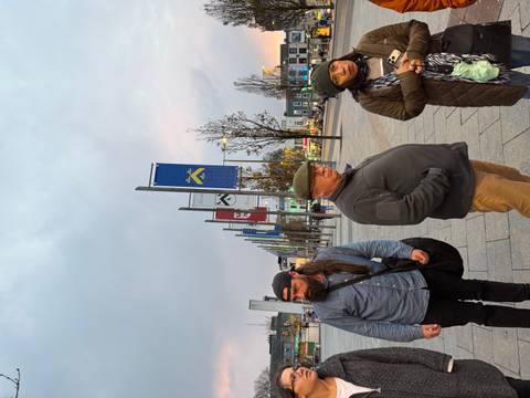 Group of people standing in a town square, flags and buildings in the background.