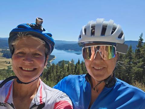 Two cyclists posing with a scenic lake and forest in the background.