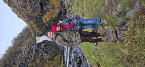 Couple standing in front of a waterfall amid rocky hills.