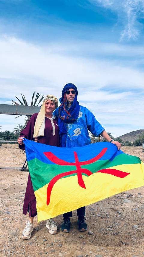 Two people holding a Berber flag with palm trees in background.