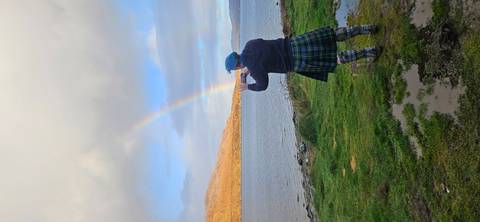      Man in a kilt taking a photo of a rainbow by a lake.
  
