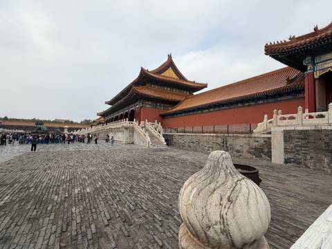 A large historical complex with traditional Chinese architecture and groups of people on the stone pavement. The buildings have intricate roofs with golden tiles and red walls.