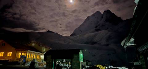 Night scene with a snowy mountain and buildings beneath a cloudy sky and moon.