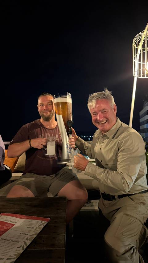       Two men enjoying large glasses of beer, smiling at a rooftop bar at night.
  