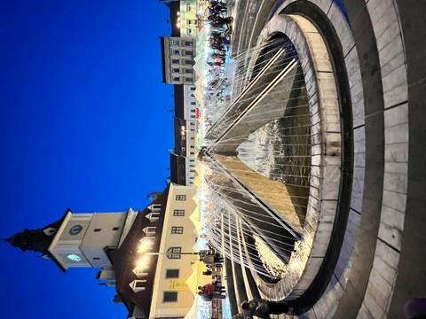       Fountain in a square with city buildings at night.
  