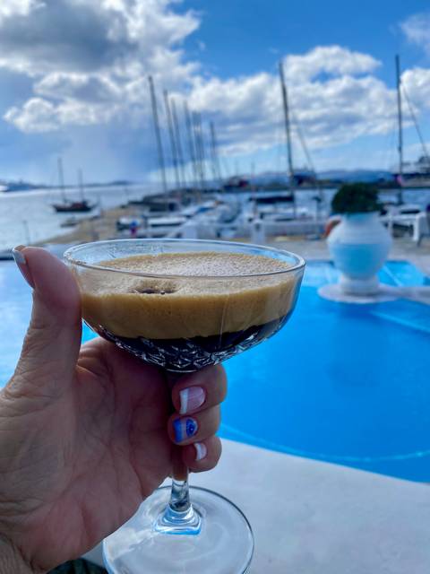       A person holding a glass of espresso martini by a swimming pool with a sea view.
  
