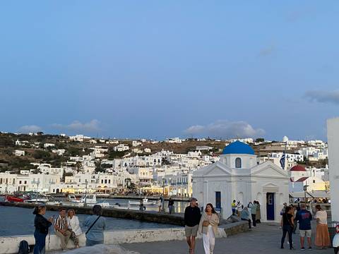       White chapel with blue dome by the sea with people walking nearby.
  
