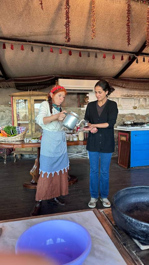 A woman pouring tea for another in a quaint interior setting with traditional decor.