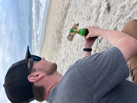 Man on a beach holding a drink with a dog lying in the background.