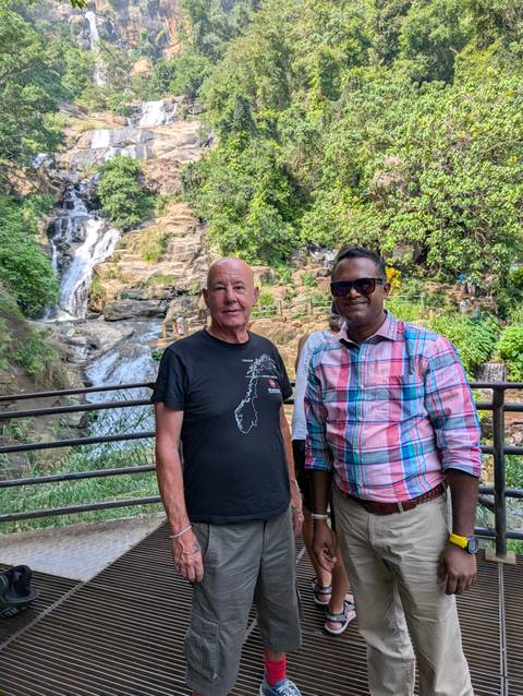 Two men posing near a waterfall and lush greenery.