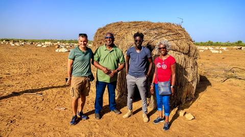 Four people standing in front of a thatched hut in a sandy landscape.