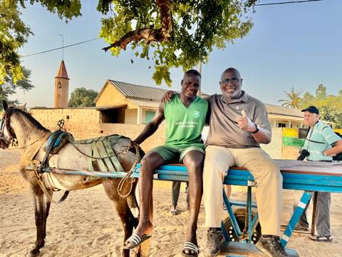 Two men sitting on a wooden cart with a horse in the background.