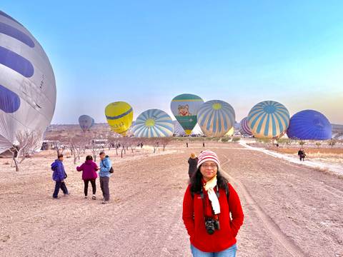 Person standing among people with hot air balloons in the background.