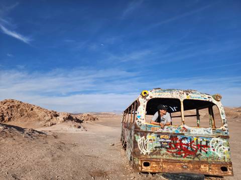       Person peering out of a colorful, graffitied bus in a desert.
  
