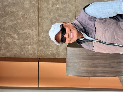       A man wearing sunglasses and a cap smiling in an outdoor area.
  